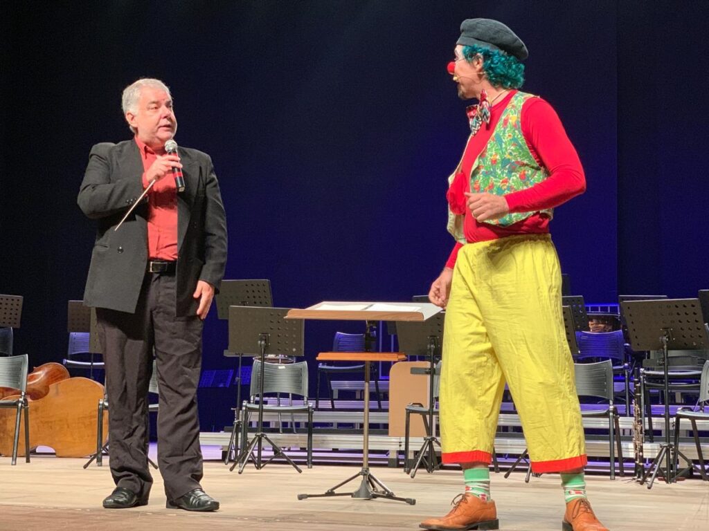 A imagem mostra dois homens em um palco com fundo azul escuro. O homem à esquerda é mais velho, com cabelo grisalho, vestindo um blazer preto sobre uma camisa vermelha, calça escura e sapatos pretos. Ele segura um microfone na mão direita e uma batuta de maestro na mão esquerda, parecendo estar falando. O homem à direita está vestido de palhaço, com cabelo verde e encaracolado, nariz vermelho, boina preta, colete colorido sobre uma camisa vermelha, gravata borboleta vermelha e verde, calças amarelas com detalhes vermelhos, meias listradas verdes e brancas e sapatos marrons. Ele olha para o homem à esquerda, com uma das mãos no peito. Ao fundo, há suportes para partituras, cadeiras e um instrumento de cordas (possivelmente um violoncelo). A luz está focada nos dois homens, destacando-os no palco, sugerindo um momento de apresentação ou ensaio.