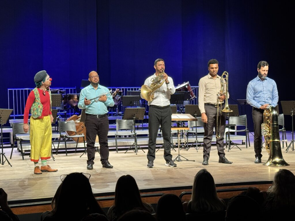 A imagem mostra uma apresentação no palco com cinco pessoas e vários instrumentos musicais. À esquerda, há um palhaço vestido com uma camisa vermelha, colete colorido, calças amarelas, boné preto, nariz vermelho e maquiagem branca no rosto. Os outros quatro indivíduos são músicos, vestidos de forma mais formal, com camisas e calças escuras. Eles seguram instrumentos de sopro: trompete, trompa, trombone e tuba. O palco é de madeira e, ao fundo, há várias cadeiras, suportes para partituras e outros instrumentos, como um contrabaixo e uma bateria, indicando que pode ser um ensaio ou concerto. A iluminação destaca os músicos, enquanto o fundo permanece escuro. Na parte inferior da imagem, é possível ver a silhueta da plateia assistindo à apresentação.