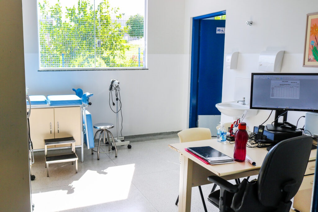 A imagem mostra uma sala de exame médico, organizada e limpa. À esquerda, há uma mesa de exame com estofado azul, equipada com apoios para as pernas e um pequeno degrau ao lado. Um banquinho giratório de metal está próximo à mesa. Na parede, uma janela grande permite a entrada de luz natural, que ilumina o ambiente e cria sombras no chão. À direita, há uma escrivaninha com um computador exibindo uma planilha, um telefone, uma garrafa vermelha e alguns materiais de escritório. Na parede próxima à escrivaninha, há uma pia com torneira e acima dela um quadro com uma imagem colorida de flores. No fundo da sala, uma porta azul com símbolos universais indica um banheiro acessível. O ambiente transmite uma sensação de profissionalismo e cuidado, típico de consultórios médicos.