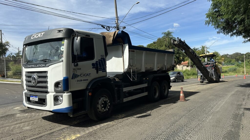 A imagem mostra uma cena de obras em uma via urbana durante o dia, com céu azul e algumas nuvens. No centro da imagem, há um caminhão branco da marca Volkswagen, modelo Constellation, com detalhes em azul e o logotipo "CICAT" visível na lateral e na frente do veículo. O caminhão está posicionado em uma rua que está sendo pavimentada ou reformada. Ao lado direito do caminhão, há uma máquina de fresagem de asfalto em operação, que está removendo a camada antiga da via. Um trabalhador usando colete amarelo de segurança está próximo à máquina, provavelmente controlando-a. Vários cones de sinalização laranja estão posicionados ao longo da rua para delimitar a área em obras. Ao fundo, é possível ver árvores verdes e algumas casas, indicando que a obra está ocorrendo em uma área residencial ou urbana. O chão apresenta marcas típicas de fresagem, com a superfície da via parcialmente removida e irregular. A iluminação natural do sol destaca os detalhes dos equipamentos e da rua em reforma.