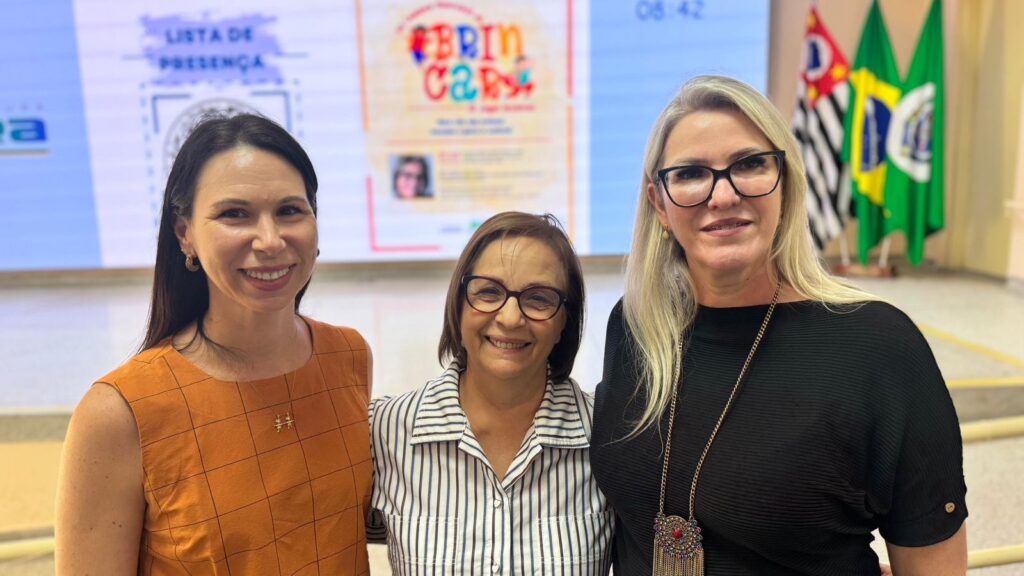 A imagem mostra três mulheres sorridentes posando para a foto em um ambiente interno que parece ser um auditório ou sala de conferências. Elas estão em pé, próximas uma da outra, transmitindo uma sensação de amizade ou parceria. A mulher à esquerda tem cabelo longo e escuro, veste uma blusa sem mangas laranja com padrão quadriculado e usa um colar dourado. A mulher no centro tem cabelo curto e castanho, usa óculos de armação preta e uma camisa listrada azul e branca. A mulher à direita tem cabelo loiro e longo, também usa óculos de armação preta, uma blusa preta texturizada e um colar dourado com um pingente grande. Ao fundo, há uma tela grande com a inscrição "LISTA DE PRESENÇA" e o logo colorido com a palavra "BRINCAR". Também são visíveis as bandeiras do Brasil e do estado de São Paulo, indicando que o evento provavelmente está acontecendo no Brasil. O ambiente é bem iluminado e parece formal, sugerindo que as mulheres participam de uma reunião, palestra ou evento oficial.