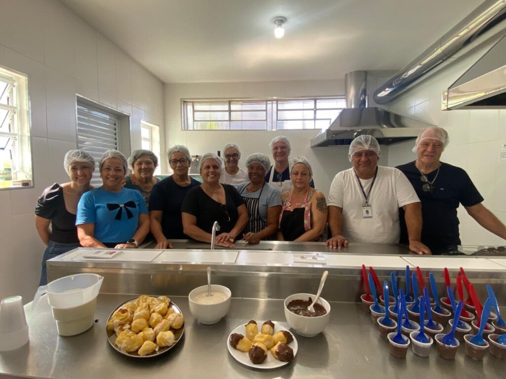 A imagem mostra um grupo de dez pessoas, sendo sete mulheres e três homens, posando para uma foto em uma cozinha comercial ou área de preparo de alimentos. Todos estão usando toucas de cabelo, indicando que estão envolvidos no manuseio de comida. Eles estão atrás de um balcão de aço inoxidável, onde há diversos alimentos e utensílios para servir. Na frente, há pratos com pequenos doces, alguns cobertos com chocolate, uma tigela com um creme branco, e vários copinhos com uma sobremesa escura, possivelmente mousse ou pudim de chocolate, cada um com uma colher pequena. O ambiente é bem iluminado pela luz natural que entra pelas janelas à esquerda, e o espaço possui paredes e piso revestidos de azulejos, além de equipamentos de cozinha em aço inox. A atmosfera parece alegre e colaborativa, com a maioria das pessoas sorrindo para a câmera.