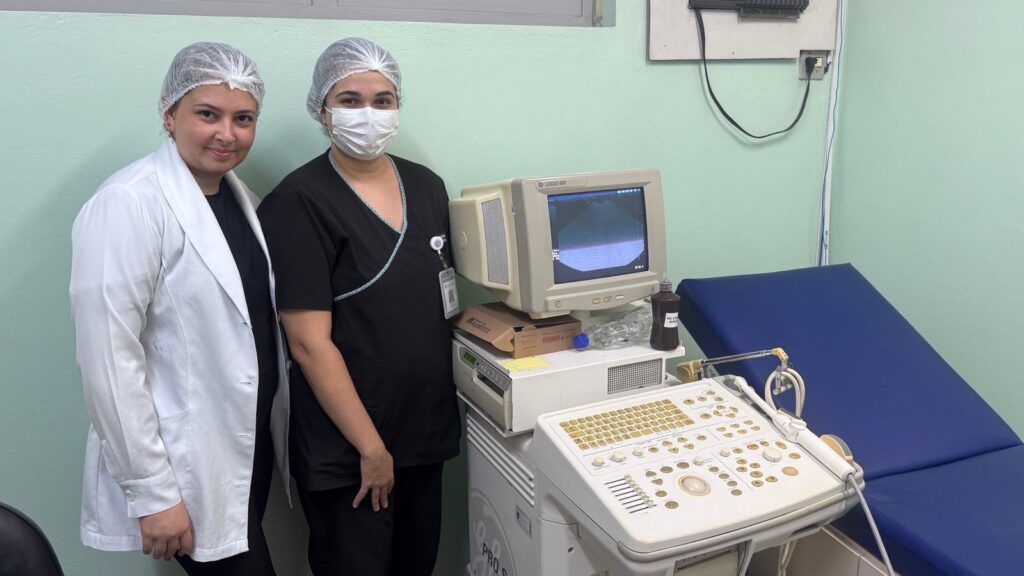 A imagem mostra duas mulheres em um ambiente clínico, possivelmente uma sala de exames médicos. Ambas usam toucas descartáveis para cabelo, e uma delas também está usando máscara cirúrgica, sugerindo um ambiente de higiene controlada. A mulher à esquerda veste um jaleco branco sobre uma blusa preta, enquanto a mulher à direita está com uniforme preto, típico de profissionais da área de saúde. Entre elas, há um aparelho de ultrassom da marca GE, modelo LOGIQ 400, com uma tela exibindo uma imagem de ultrassom. O equipamento possui um painel de controle cheio de botões e um teclado. Ao lado do aparelho, há uma maca azul, usada para acomodar pacientes durante exames. Na prateleira acima do aparelho, há frascos de gel condutor para ultrassom, um deles identificado parcialmente como "GEL CO... 240ML". O ambiente tem paredes verdes claras e iluminação clara, típica de clínicas ou hospitais. A atmosfera é profissional e calma, sugerindo que as mulheres podem ser profissionais de saúde, possivelmente preparadas para realizar ou já tendo realizado um exame de ultrassom.