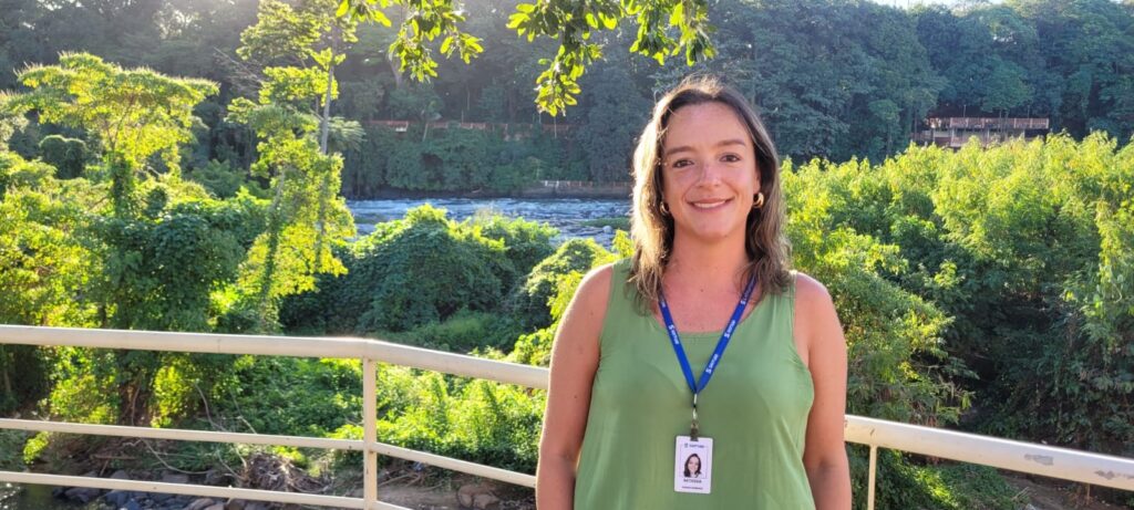 A imagem mostra uma mulher sorridente em um ambiente ao ar livre, cercada por uma paisagem natural exuberante. Ela está posicionada no centro-direito da foto, usando um top verde claro sem mangas e brincos de argola grandes. Ela tem cabelo castanho na altura dos ombros e usa um crachá azul com seu nome, "Pamela Rodriguez", e a palavra "NATASSIA" visível. Ao fundo, há muitas árvores verdes e um rio, com luz do sol suave iluminando a cena, criando um efeito de luz e sombra nas folhas próximas. A foto transmite uma sensação de tranquilidade e conexão com a natureza, com um cenário que parece ser um parque ou reserva natural.