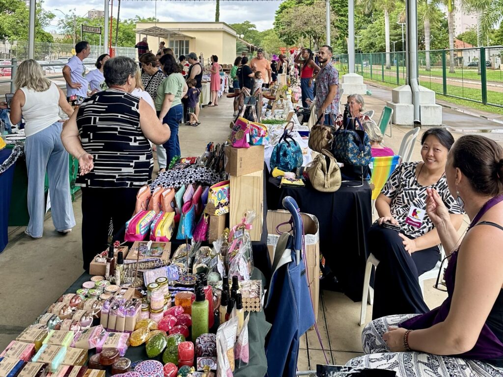 A imagem mostra uma feira ao ar livre, provavelmente um evento de artesanato ou mercado local, com várias barracas organizadas sob uma estrutura coberta. No primeiro plano, há uma mesa longa coberta com toalha escura, repleta de produtos artesanais, como sabonetes, cosméticos e bolsas coloridas. Os produtos estão organizados de forma atraente, alguns embalados, outros expostos individualmente. Diversas pessoas circulam pelo local, algumas observando os produtos, outras conversando entre si. Elas estão vestidas de maneira casual e parecem interessadas nas mercadorias expostas. Um dos indivíduos usa um crachá com a palavra "EXPOSITOR", indicando que é um vendedor ou responsável por uma das barracas. O ambiente é aberto, com árvores e construções ao fundo, sugerindo que a feira acontece em um parque ou área urbana com espaço aberto. O céu está nublado, o que proporciona uma iluminação suave e difusa. Na parte de trás, há uma placa com a palavra "PIRACICABA", indicando a provável localização do evento, que é uma cidade no Brasil. A atmosfera geral da imagem é de um evento comunitário, focado em produtos artesanais e contato direto entre vendedores e compradores.