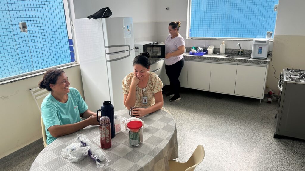 A imagem mostra uma cozinha ou copa iluminada, onde três mulheres estão presentes. Duas delas estão sentadas em uma mesa redonda coberta por uma toalha cinza com estampa geométrica. A mulher à esquerda veste uma camiseta azul-turquesa e sorri, olhando para a companheira à direita, que usa uma blusa de estampa floral e segura uma caneca preta, também sorrindo. Sobre a mesa há dois garrafas térmicas coloridas (uma azul e outra vermelha), um pote transparente com tampa vermelha contendo algum alimento e alguns itens embalados em plástico. Ao fundo, uma terceira mulher, vestida com camiseta rosa clara e calça preta, está de pé próxima a uma bancada de cozinha, aparentemente utilizando um micro-ondas. A cozinha é equipada com geladeira branca, armários brancos com tampo de granito, pia, purificador de água e fogão. Duas janelas grandes com grade azul permitem a entrada de luz natural. O ambiente tem um aspecto acolhedor e informal, sugerindo um momento de pausa ou convivência em um local de trabalho ou residencial.