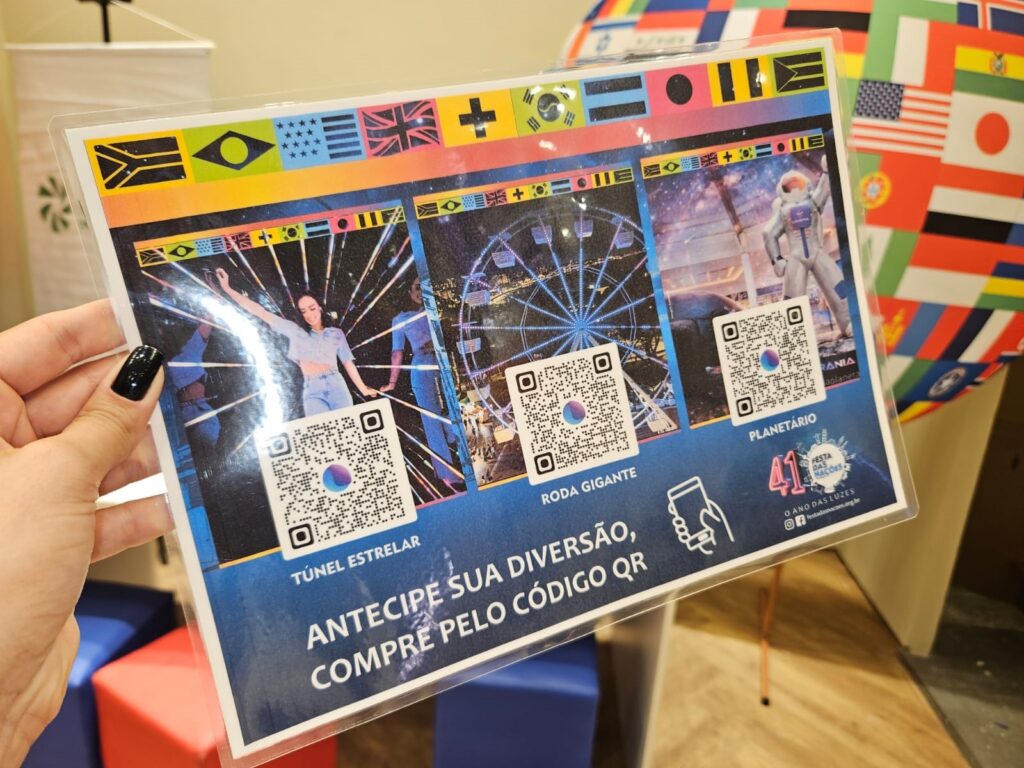 A imagem mostra uma mão segurando um folheto laminado, colorido e com design vibrante. O folheto tem um fundo estrelado e uma faixa superior com bandeiras de vários países, sugerindo um tema internacional. Ele está dividido em três seções principais, cada uma com uma imagem e um código QR abaixo: À esquerda, uma mulher dançando em um ambiente com luzes coloridas, representando o "TÚNEL ESTRELAR". No centro, uma roda-gigante iluminada à noite, chamada "RODA GIGANTE". À direita, um astronauta em um cenário que parece um planetário, identificado como "PLANETÁRIO". Abaixo dessas imagens, há um texto que diz: "ANTECIPE SUA DIVERSÃO, COMPRE PELO CÓDIGO QR". Na parte inferior direita do folheto, está o logo da "41ª FESTA DAS NAÇÕES" com o slogan "O ANO DAS LUZES" e o site "festadasnacoes.org.br". A mão que segura o folheto tem unhas pintadas de preto, e o fundo da imagem está desfocado, indicando que a foto foi tirada com foco no folheto. A imagem transmite uma atmosfera festiva e convida o público a participar do evento cultural, comprando ingressos antecipadamente via QR code.