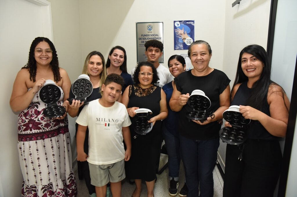 A imagem mostra um grupo de nove pessoas reunidas em um ambiente interno, provavelmente uma sala de eventos ou reunião. Entre elas, há sete mulheres, um jovem e um menino. Cinco das mulheres estão segurando aparelhos pequenos, brancos, que parecem ser máquinas portáteis para fazer donuts, abertas para mostrar as cavidades onde os donuts são preparados. O grupo está alinhado em duas fileiras, com os mais baixos e o menino na frente, e as mulheres e o jovem atrás. Todos estão sorrindo, transmitindo uma sensação de celebração ou conquista. O menino, que está na frente, usa uma camiseta branca com a inscrição "MILANO ITALIA". Ao fundo, há uma parede branca com uma placa comemorativa em metal e um cartaz azul. A placa tem inscrições em português, indicando que o local está ligado ao "Fundo Social de Solidariedade de Piracicaba", sugerindo que a foto foi tirada em uma entidade ou evento social na cidade de Piracicaba. A iluminação é clara e uniforme, destacando bem as pessoas e os objetos que elas seguram. A composição da foto transmite um clima de união, aprendizado ou entrega de algum projeto ou produto, possivelmente relacionado à fabricação de donuts, dado os aparelhos mostrados.