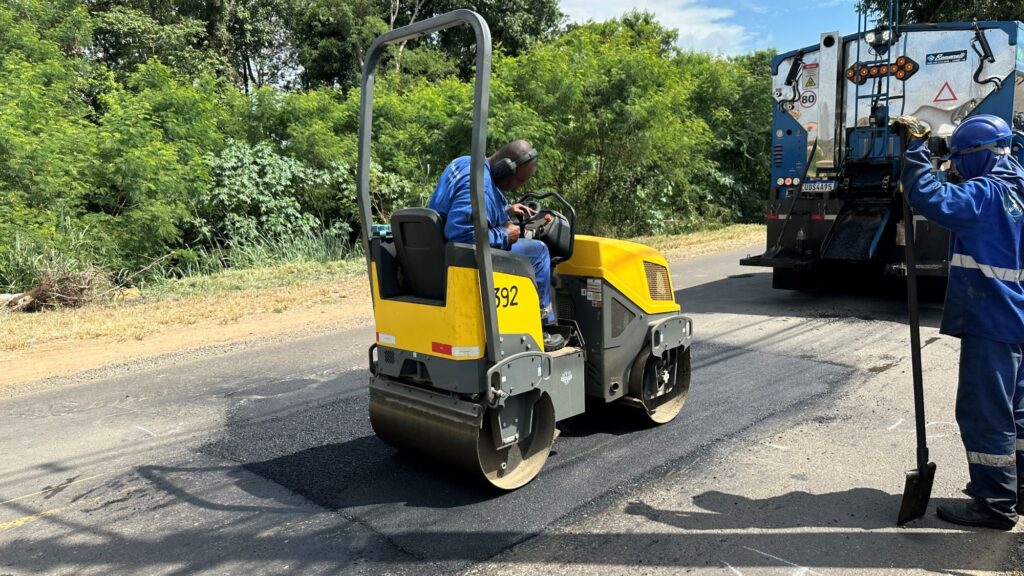 A imagem mostra uma cena de manutenção rodoviária em uma estrada de duas pistas durante um dia ensolarado. No centro da imagem, há uma compactadora de asfalto amarela e cinza, sendo operada por um homem vestido com uniforme azul e protetores auriculares. A compactadora está sobre uma área de asfalto recém-aplicado, que é mais escura e visivelmente diferente do restante da pista. À direita da compactadora, há outro trabalhador também com uniforme azul e capacete azul, segurando uma ferramenta longa, possivelmente uma pá ou um ancinho, provavelmente para auxiliar no espalhamento do asfalto. Mais ao fundo, na mesma faixa da estrada, está um caminhão distribuidor de asfalto azul e branco, equipado com diversos sinais de segurança, como uma placa indicando velocidade máxima de 80 km/h e outras sinalizações refletivas. Ao fundo, há vegetação densa, com muitas árvores verdes, indicando que a estrada está em uma área mais rural ou com bastante natureza ao redor. A iluminação é forte, gerando sombras nítidas no asfalto e destacando os detalhes da cena. No geral, a imagem mostra o processo de reparo ou pavimentação da estrada, com os trabalhadores em ação e os equipamentos necessários para a tarefa.