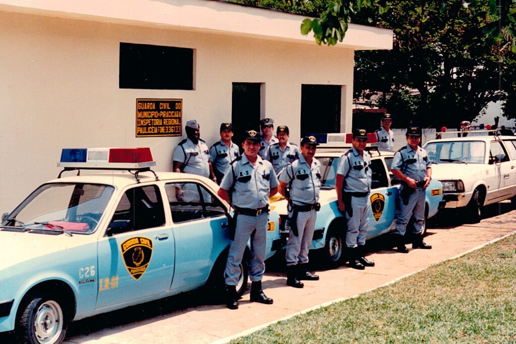 A imagem mostra um grupo de nove homens uniformizados, provavelmente membros da Guarda Civil do município de Piracicaba, em pé em frente a um prédio claro com janelas retangulares escuras. Eles estão alinhados em duas fileiras e parecem estar posando para a foto. Todos vestem uniformes cinza com detalhes escuros, incluindo bonés, cintos e distintivos. Ao lado deles, há dois carros de polícia com cores predominantes azul claro e branco, ambos com sirenes vermelhas e azuis no teto. Um dos carros exibe claramente a inscrição "GUARDA CIVIL" e um emblema circular com um escudo. Na parede do prédio, há uma placa com o texto "GUARDA CIVIL DO MUNICIPIO-PIRACICABA INSPETORIA REGIONAL POLICETAFONE:336133", indicando que a cena se passa em uma inspetoria regional da Guarda Civil em Piracicaba. A iluminação é natural, provavelmente durante o dia, e o cenário transmite uma sensação oficial e institucional. A foto parece ser antiga, com cores um pouco desbotadas e um leve granulado típico de imagens de décadas passadas.