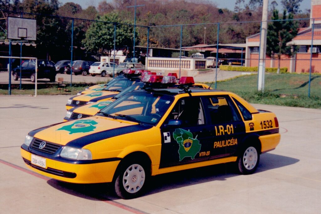 A imagem mostra uma linha de três carros da Volkswagen, provavelmente veículos policiais, estacionados em um piso de asfalto. Os carros são predominantemente amarelos com detalhes em azul escuro, incluindo uma faixa que percorre a lateral e o capô. Cada veículo tem um emblema verde no capô e na lateral, e há uma barra de luz vermelha e branca no teto, típica de viaturas policiais. Na frente do primeiro carro, há uma placa com a inscrição "I.R-01" e outras marcações, como "PAULICEIA", "VTR-20", "DBA 4594" e o número "1532". Ao fundo, é possível ver uma cerca de arame farpado, algumas árvores e edifícios que parecem fazer parte de um complexo institucional, possivelmente uma delegacia ou centro de treinamento policial. Também há uma cesta de basquete à esquerda, indicando que o local pode ter uma área de lazer. A imagem foi capturada em um dia claro, com boa iluminação natural, e a perspectiva é levemente baixa, destacando os veículos em primeiro plano. Os carros estão alinhados de forma ordenada, transmitindo uma sensação de organização e prontidão.