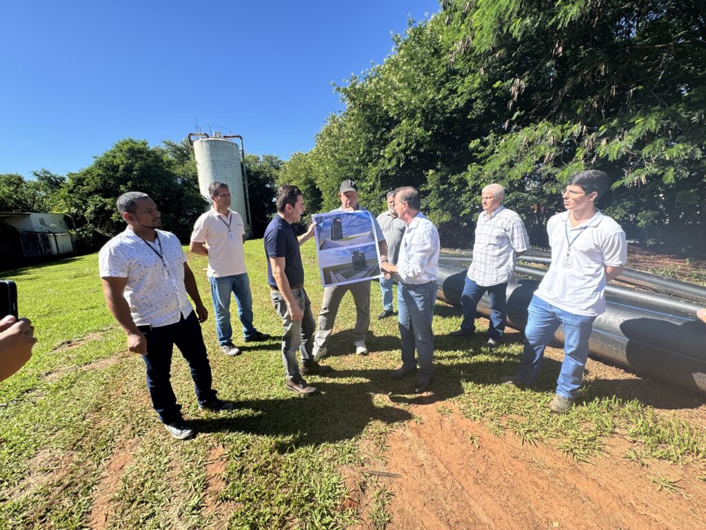 A imagem mostra um grupo de oito homens reunidos ao ar livre, em um ambiente que parece ser semi-rural ou industrial. O foco principal está em um homem no centro, que segura um documento impresso com duas imagens aéreas ou renderizações de grandes estruturas cilíndricas escuras, possivelmente tanques de água ou equipamentos industriais. Ele está gesticulando em direção ao documento, aparentemente explicando algo aos outros homens. O grupo é composto por pessoas de diferentes idades, vestindo roupas casuais, como camisas e calças jeans. O cenário ao redor inclui uma área com grama, caminhos de terra, árvores verdes ao fundo e uma torre de concreto alta e desgastada à esquerda. Também é possível ver grandes tubos pretos à direita da imagem. A iluminação é forte, indicando que a foto foi tirada durante o dia, com sombras bem definidas. A composição da foto é de plano médio, capturando a interação entre as pessoas e o ambiente ao redor.