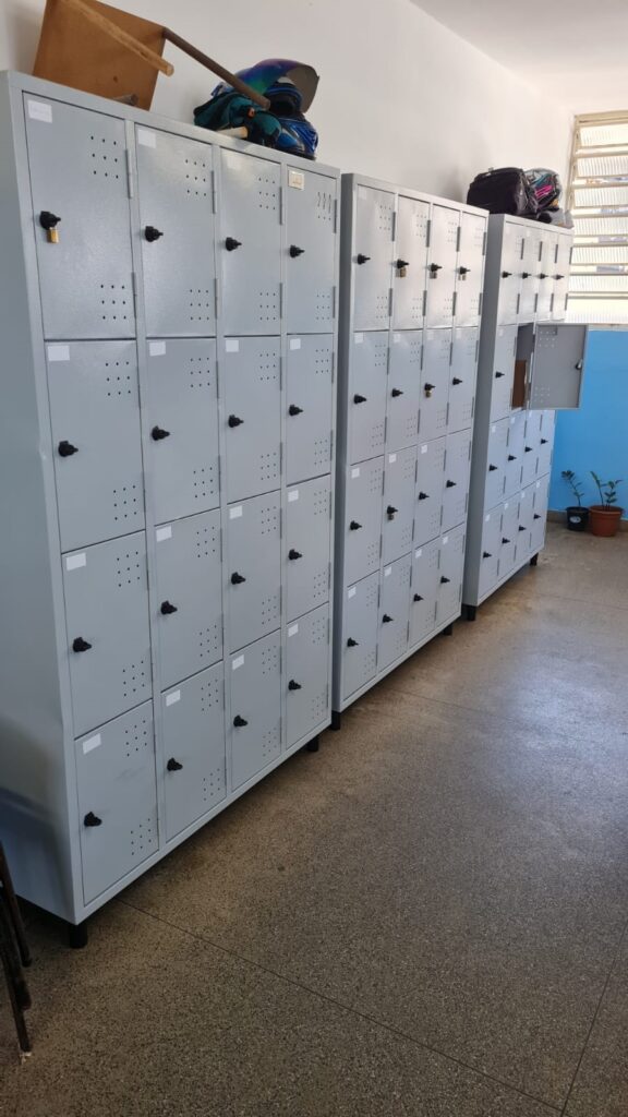 A imagem mostra três conjuntos grandes de armários metálicos cinza claro, dispostos em linha contra uma parede branca em um ambiente interno, possivelmente um corredor ou uma sala. O conjunto à esquerda é composto por 20 portas organizadas em uma grade 4x5, o do meio também tem 20 portas (4x5), e o da direita é mais estreito, com 15 portas em uma grade 3x5. Cada porta possui um puxador preto, pequenos furos de ventilação organizados em grupos de nove e um espaço para etiqueta branca no canto superior esquerdo. Algumas portas estão trancadas com cadeados, enquanto uma porta na unidade da direita está aberta, mostrando o interior escuro do armário. No topo do conjunto da esquerda há uma tábua de madeira e um capacete de motociclista azul com detalhes pretos. No armário da direita, há uma mochila preta sobre ele. O chão é de um material tipo granulado, em tons de cinza e bege. Ao fundo, a parede branca encontra uma parede azul clara onde há duas pequenas plantas em vasos próximos a uma janela com persianas horizontais. A iluminação é clara e uniforme, provavelmente vinda de luz natural pela janela ou de luminárias no teto. A imagem transmite uma sensação de organização e simplicidade, com um padrão repetitivo das portas dos armários.