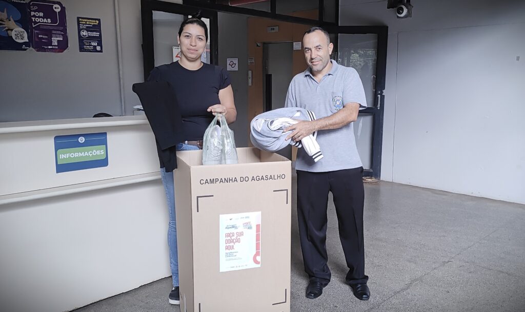A imagem mostra duas pessoas, um homem e uma mulher, participando de uma campanha de doação de roupas de inverno, chamada "Campanha do Agasalho". Eles estão ao lado de uma grande caixa de papelão, onde as doações são colocadas. A caixa está identificada com a frase "CAMPANHA DO AGASALHO" e "FAÇA SUA DOAÇÃO AQUI". A mulher, vestindo camiseta preta e calça jeans azul, segura um saco plástico com sapatos e uma peça de roupa preta. O homem, de camiseta cinza clara e calça preta, segura uma blusa cinza dobrada. Ambos estão sorrindo para a câmera. Ao fundo, há uma mesa de atendimento com a palavra "INFORMAÇÕES" e várias placas nas paredes, incluindo um cartaz relacionado a Narcóticos Anônimos e outro com a frase "SP POR TODAS". O ambiente parece ser uma recepção ou área de entrada de uma instituição. A iluminação é clara, provavelmente natural ou fluorescente, e o local está bem organizado. A cena transmite uma mensagem de solidariedade e incentivo à doação para ajudar pessoas em situação de vulnerabilidade durante o inverno.
