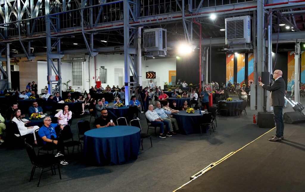 A imagem mostra um evento realizado em um salão amplo com estrutura industrial, visível pelas vigas metálicas e tubulações expostas no teto. O ambiente está iluminado por luzes fortes que criam um contraste marcante entre áreas claras e sombras, conferindo um clima dramático à cena. À direita, um homem calvo, vestido com traje formal escuro, está em pé, segurando um microfone e fazendo gestos com a mão, sugerindo que está discursando ou apresentando algo para a plateia. A audiência, composta por cerca de 50 a 70 pessoas de diferentes idades e gêneros, está sentada em mesas redondas cobertas por toalhas azuis escuras, algumas com pequenos arranjos florais. A maioria das pessoas está atenta ao palestrante, enquanto algumas conversam entre si ou olham para seus celulares. O espaço é amplo, com piso escuro e várias faixas ou banners pendurados ao fundo, que parecem conter o nome "ECOBRISA" e a palavra "SAÍDA", além de referências a "Salão de Reunião" e "Campinas". O cenário sugere uma reunião corporativa ou conferência, com um clima profissional e organizado, destacando o palestrante como o centro das atenções. A iluminação e a composição da imagem valorizam a interação entre o orador e o público presente.