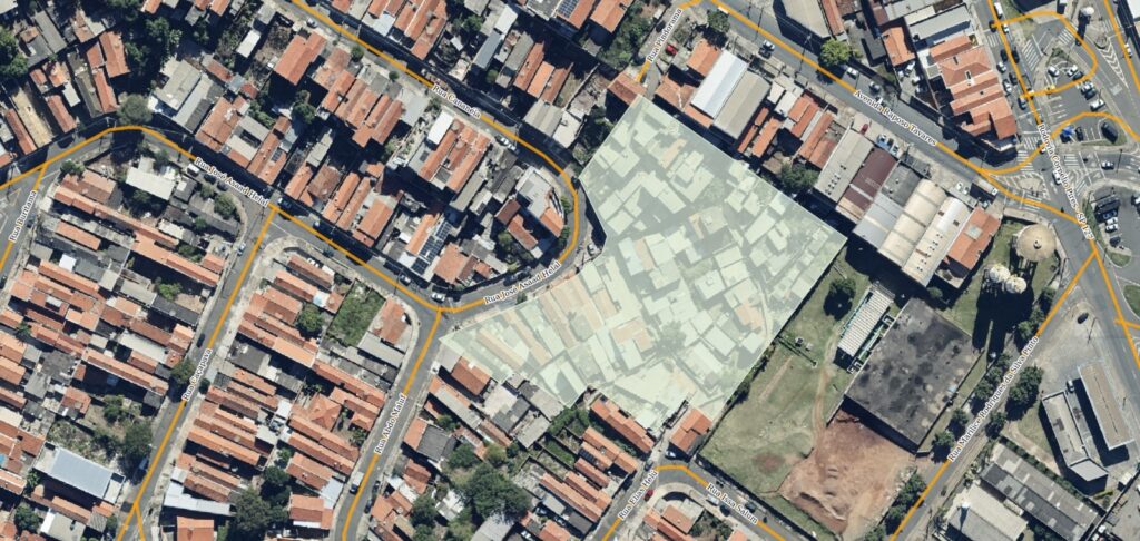 A imagem mostra uma vista aérea de uma área urbana densamente povoada, provavelmente um bairro residencial com algumas construções comerciais ou industriais. As casas têm telhados predominantemente vermelhos, típicos de áreas residenciais. As ruas são estreitas e sinuosas, com algumas avenidas maiores e uma rodovia visível ao fundo, identificada como Rodovia Cornélio Pires (SP-127) e Avenida Raposo Tavares. No centro da imagem, há uma área destacada que parece ser uma favela ou um conjunto de habitações informais, com construções muito próximas e irregulares. Ao redor, há ruas nomeadas, como Rua Buritama, Rua Caçapava, Rua José Asaad Helal, Rua Abdo Maluf, entre outras. Também é possível ver algumas áreas com vegetação e árvores espalhadas, além de algumas construções maiores que parecem ser indústrias ou galpões, incluindo uma com uma torre de água. Há ainda um campo aberto ou quadra esportiva em terra. A imagem apresenta um contraste entre a organização das ruas e construções formais e a densidade e desorganização do conjunto habitacional informal no centro. A iluminação indica que a foto foi tirada durante o dia, com sombras projetadas pelas construções.