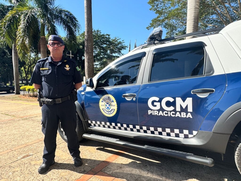 A imagem mostra um agente da Guarda Civil Metropolitana (GCM) de Piracicaba em pé ao lado de uma viatura oficial. O agente está vestido com uniforme azul escuro, incluindo uma boina e um cinto de utilidades, que provavelmente contém equipamentos de trabalho. Ele está olhando para a câmera com uma expressão séria. A viatura, uma SUV com pintura azul e branca, tem o emblema da Guarda Civil Metropolitana de Piracicaba estampado na porta e as siglas "GCM PIRACICABA" visíveis. Também aparece o número "153", que é o telefone de emergência da guarda. Ao fundo, há árvores de palmeira e um céu azul claro, indicando que a foto foi tirada em um dia ensolarado. A cena transmite uma sensação de autoridade e segurança pública, com o agente e a viatura prontos para atender a comunidade local.