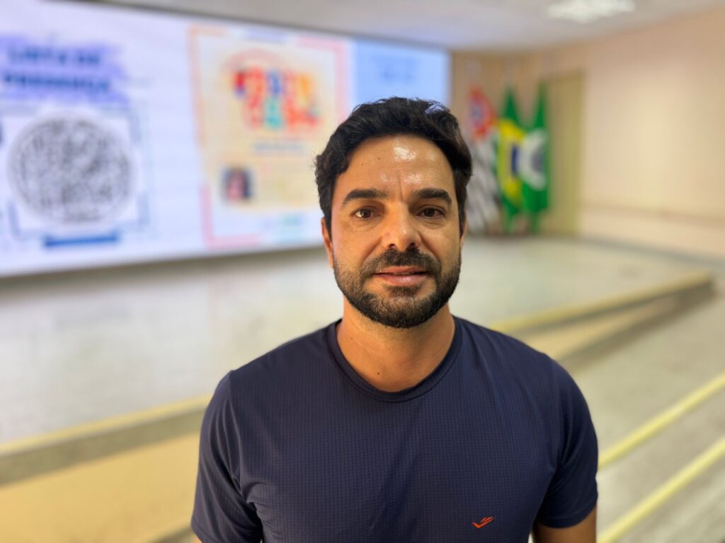 A imagem mostra um homem de meia-idade, com cabelo castanho escuro e ondulado, barba curta e bem cuidada. Ele veste uma camiseta azul marinho com um pequeno logo laranja no peito. O fundo está desfocado, mas é possível perceber que ele está em um ambiente interno, possivelmente um auditório ou sala de conferências. Atrás dele, à direita, há três bandeiras: a bandeira do estado de São Paulo, a bandeira do Brasil e outra bandeira verde. À esquerda, há uma tela grande com gráficos e textos, parcialmente visíveis, incluindo um código QR. A iluminação é clara e uniforme, destacando o rosto do homem, que tem uma expressão neutra.