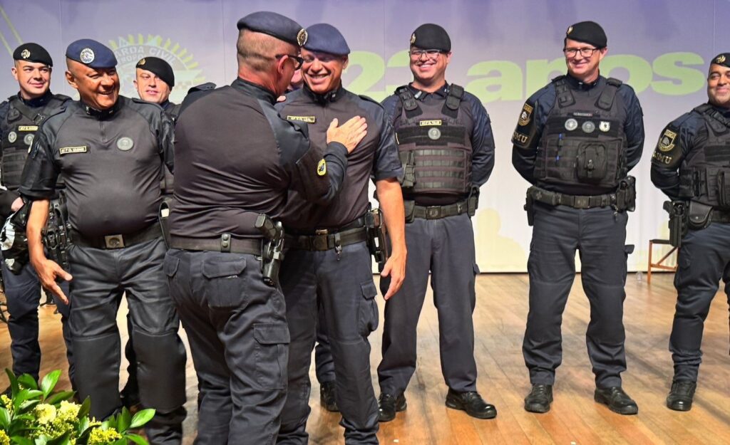 A imagem mostra um grupo de homens fardados, provavelmente integrantes da Guarda Civil, reunidos em um evento oficial. Dois deles estão em destaque no centro da imagem: um homem está sorrindo enquanto coloca a mão no ombro do outro, que também sorri. Eles parecem estar celebrando ou reconhecendo algo, demonstrando um momento de camaradagem e respeito. Os homens vestem uniformes escuros, com coletes táticos e insígnias, indicando que são agentes de segurança ou policiais. Ao fundo, há uma parede com um grande símbolo e textos que incluem "GUARDA CIVIL", "KROPOS" e "20 ANOS", sugerindo que a ocasião pode ser uma comemoração dos 20 anos dessa organização ou de uma unidade específica. A iluminação é clara e uniforme, destacando bem os rostos e os detalhes dos uniformes. A composição da foto é formal, com os homens alinhados e focados na interação central. A atmosfera geral transmite profissionalismo, respeito e celebração.