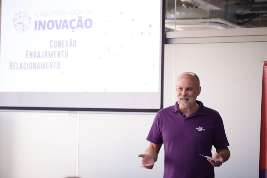 A imagem mostra um homem apresentando em frente a uma tela de projeção. Ele está vestido com uma camiseta polo roxa que tem o logo do SEBRAE, indicando que a apresentação está relacionada a essa instituição. O homem parece estar falando e gesticulando com a mão direita, enquanto segura um pedaço de papel na mão esquerda, transmitindo uma postura confiante e engajada. Na tela de projeção, há um texto em português que diz: "ECOSSISTEMA LOCAL DE INOVAÇÃO" em destaque, seguido pelas palavras "CONEXÃO", "ENGAJAMENTO" e "RELACIONAMENTO", sugerindo que o tema da apresentação está relacionado à criação e fortalecimento de redes e parcerias para inovação local. O ambiente parece ser uma sala de reunião ou auditório, com uma iluminação clara que destaca tanto o apresentador quanto a tela. A composição da imagem foca no homem e na tela, transmitindo um momento de compartilhamento de conhecimento ou treinamento.