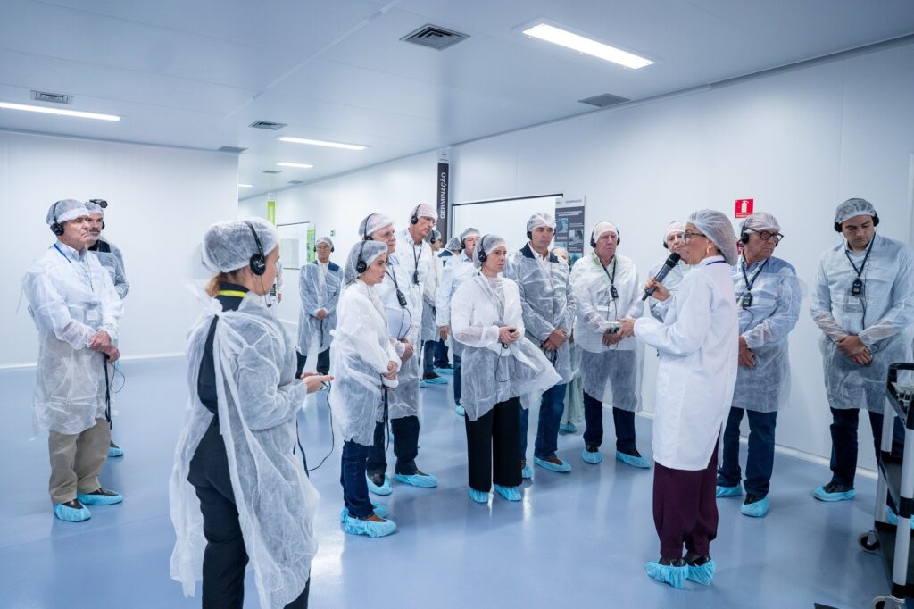 A imagem mostra um grupo de pessoas dentro de um ambiente limpo e controlado, possivelmente uma instalação de pesquisa, laboratório ou fábrica. Todos os indivíduos estão vestidos com aventais brancos, toucas para cobrir o cabelo e protetores azuis nos calçados, indicando que estão em uma área que exige alta higiene, como uma sala limpa. Eles usam fones de ouvido, sugerindo que estão participando de uma visita guiada com áudio sincronizado. À frente do grupo, uma mulher também vestida com o uniforme de proteção fala ao microfone, provavelmente a guia ou instrutora do grupo. No fundo, é possível ver paredes brancas e azuis, além de placas com a palavra "GERMINAÇÃO", indicando que o local pode estar relacionado a processos de cultivo ou desenvolvimento de plantas, sementes ou algo similar. A iluminação é forte e uniforme, típica de ambientes controlados. A imagem transmite uma atmosfera de profissionalismo, cuidado e controle rigoroso de higiene.