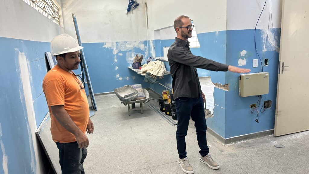 A imagem mostra um ambiente interno em processo de reforma. Dois homens estão na cena: um deles, à esquerda, é um trabalhador da construção civil, vestindo capacete branco, camiseta laranja e calça escura, olhando para a direita. O outro homem, à direita, usa óculos, tem barba curta e está vestido com uma camisa cinza escura e jeans azul, apontando para uma parede à sua frente. A parede apresenta sinais visíveis de desgaste, como pintura descascada e áreas de concreto exposto. No centro da sala, há uma carroça de mão metálica e, ao redor, diversos materiais de construção, como sacos de cimento e baldes de tinta. O chão é revestido com um piso cinza claro com pequenos pontos escuros. A luz natural entra pela janela ao fundo e pela porta aberta à direita, iluminando o ambiente. A cena transmite uma atmosfera de trabalho e planejamento na reforma do espaço.