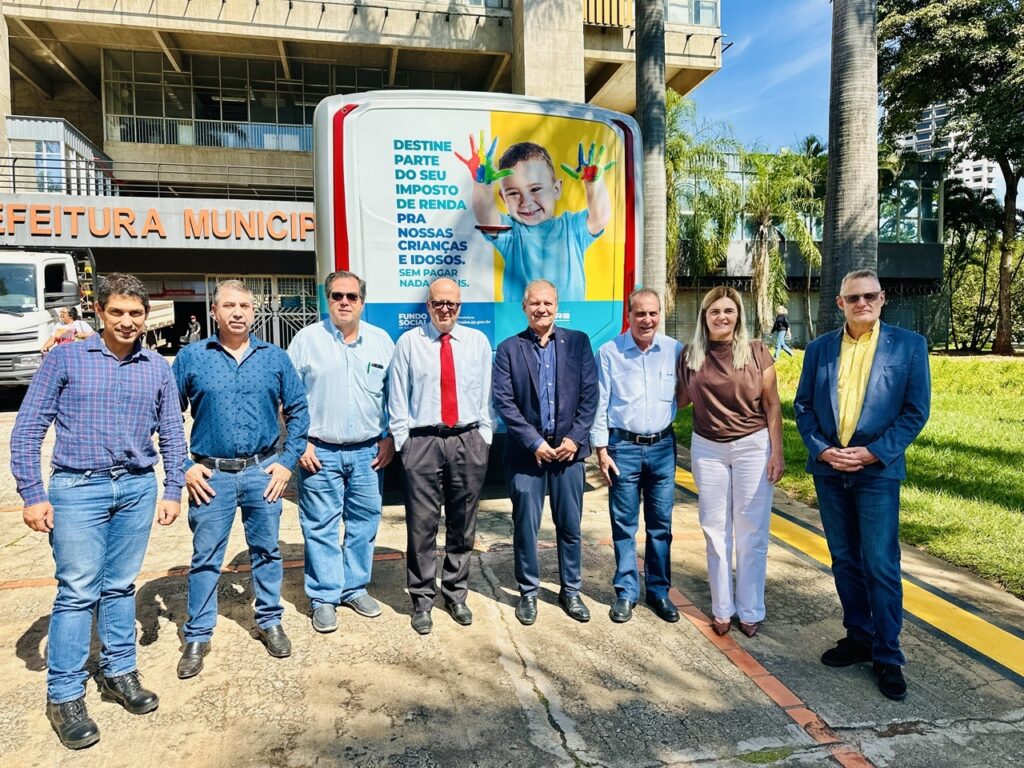 A imagem mostra um grupo de oito pessoas (seis homens e duas mulheres) posando para uma foto em frente a um prédio público, identificado como "Prefeitura Municipal". Atrás deles, há um grande painel publicitário vertical, que exibe a imagem de mãos de criança pintadas com várias cores. O texto no painel está em português e incentiva a destinação de parte do imposto de renda para ajudar crianças e idosos, sem custo para o contribuinte, mencionando o "Fundo Social". O cenário é ao ar livre, com céu claro e sol brilhando, indicando que a foto foi tirada durante o dia. Há palmeiras e árvores ao fundo, além de uma área gramada, sugerindo um espaço público bem cuidado. O grupo está alinhado em frente ao painel e ao prédio, todos olhando para a câmera, com expressões neutras ou de leve sorriso. As pessoas estão vestidas de forma casual e semiformal. O ambiente transmite uma atmosfera institucional e de campanha social, com o objetivo de conscientizar a população sobre a possibilidade de ajudar grupos vulneráveis por meio do imposto de renda. A combinação do prédio da prefeitura, o painel informativo e o grupo de pessoas sugere que se trata de uma ação oficial ou evento público.