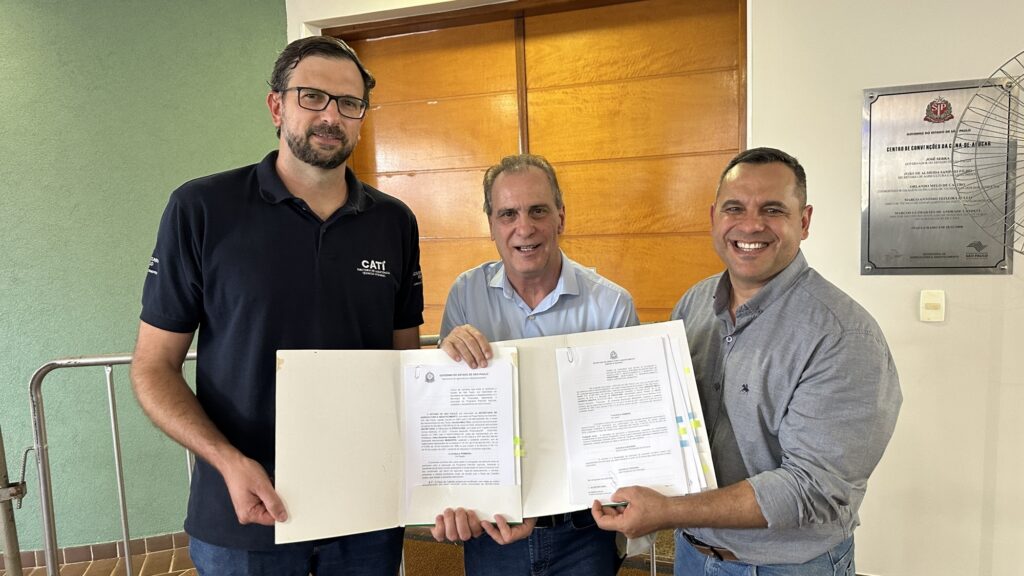 A imagem mostra três homens em um ambiente interno, posando para a foto enquanto seguram juntos uma pasta com documentos. O homem à esquerda veste uma camiseta polo azul escura com o logo "CATI" e usa óculos. O homem no centro usa uma camisa azul clara e sorri para a câmera, enquanto o homem à direita veste uma camisa cinza e também está sorrindo. Eles parecem estar em um momento formal, possivelmente relacionado à assinatura de algum acordo ou apresentação de documentos. Ao fundo, há uma parede com painéis de madeira e uma placa com texto e logotipo, sugerindo um ambiente institucional ou de escritório. A iluminação é clara, destacando os rostos e os documentos que estão segurando. A expressão dos homens transmite satisfação e profissionalismo.