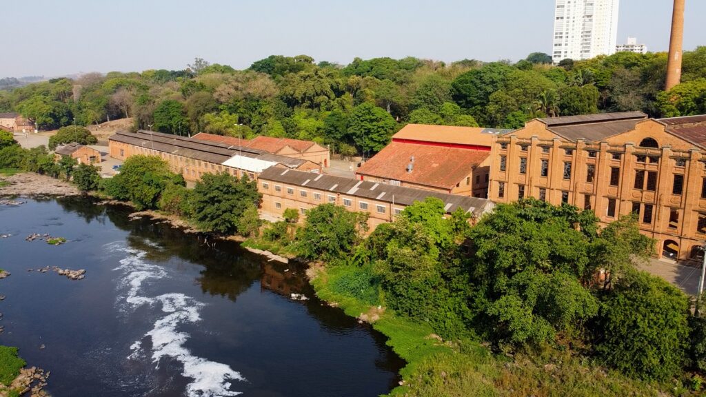 A imagem mostra uma vista aérea de um complexo industrial histórico feito de tijolos vermelhos, localizado à beira de um rio de águas escuras. O complexo é composto por vários prédios longos e baixos, com telhados de duas águas, alguns cobertos por telhas laranja e outros por um material corrugado cinza. À direita da imagem, há uma chaminé alta e esguia, também feita de tijolos vermelhos. Ao redor dos prédios, há muitas árvores e vegetação densa, que criam um contraste com a cor dos tijolos. Ao fundo, é possível ver uma área urbana com um prédio alto e moderno, branco, que destaca a mistura entre o antigo e o contemporâneo. A luz do sol é intensa, iluminando toda a cena de forma clara, sugerindo que a foto foi tirada durante o dia. O rio corre próximo ao complexo, separando-o da vegetação e criando um cenário pitoresco e histórico.
