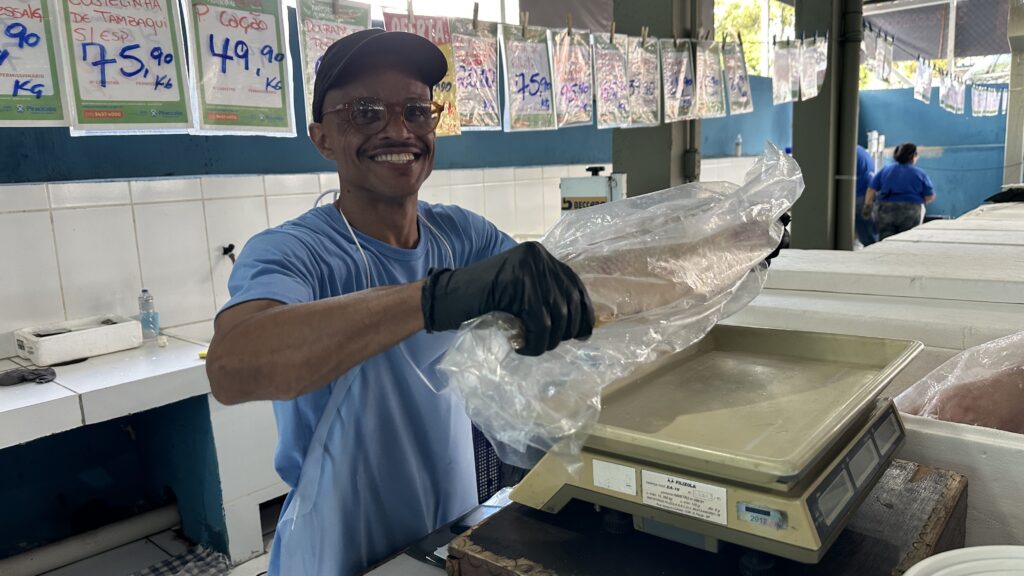 A imagem mostra um homem sorridente em um ambiente de peixaria. Ele está no centro da foto, usando boné escuro, óculos, camiseta azul clara e avental branco, além de luvas pretas. Ele segura um peixe grande embalado em plástico transparente e o coloca em uma balança digital da marca Filizola. Atrás dele, há várias placas com nomes e preços de peixes, escritos em português e em reais por quilograma, como "Costelinha de Tambaqui" a Rã 49,90/kg. O fundo é composto por paredes azuis e brancas, azulejos e caixas térmicas de isopor, típicas de um mercado de peixes. O ambiente está bem iluminado, transmitindo uma sensação de profissionalismo e simpatia.