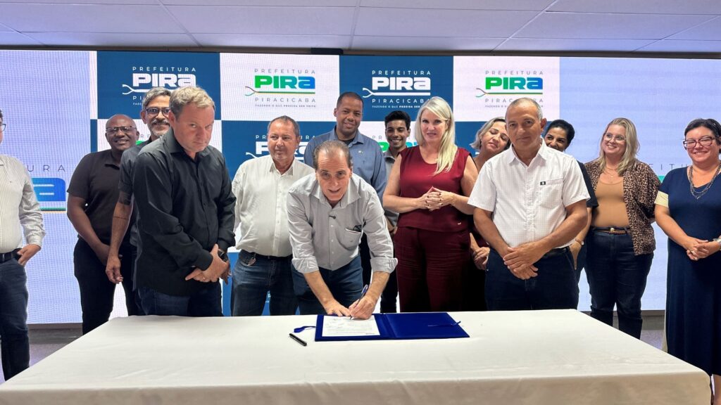 A imagem mostra uma cerimônia oficial de assinatura, possivelmente relacionada a um ato governamental na cidade de Piracicaba, Brasil. No centro da cena, um homem está curvado sobre uma mesa branca, assinando um documento colocado dentro de uma pasta azul. Ao seu redor, há um grupo de cerca de doze pessoas, homens e mulheres de diferentes idades, que observam o momento com expressões de atenção e satisfação. Eles estão vestidos de forma casual e formal, com camisas, blusas e calças. Ao fundo, há um painel azul com o logotipo e o nome da Prefeitura de Piracicaba, acompanhado do slogan “Fazendo o que precisa ser feito”, reforçando o caráter oficial e institucional do evento. A iluminação é clara e destaca todos os participantes, transmitindo um ambiente sério e solene, típico de eventos públicos e governamentais. A disposição das pessoas em semicírculo ao redor do homem que assina sugere apoio e testemunho coletivo ao ato que está sendo realizado.