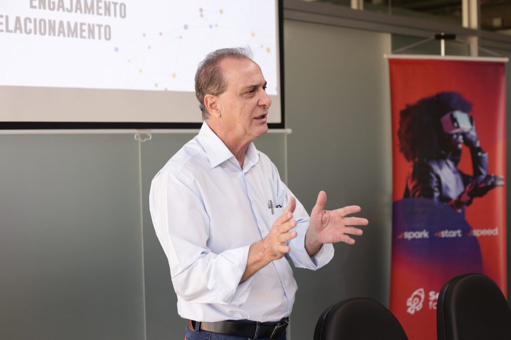 A imagem mostra um homem de meia-idade apresentando-se em um ambiente que parece ser uma conferência ou palestra. Ele está em pé, vestindo uma camisa azul clara e calças escuras, com uma caneta no bolso da camisa. O homem está gesticulando com as mãos, possivelmente enfatizando algum ponto durante sua fala. Ao fundo, há uma tela de projeção exibindo texto e gráficos, embora o conteúdo específico da projeção não esteja totalmente legível, exceto pelas palavras "ENGAJAMENTO" e "RELACIONAMENTO". À direita da imagem, há um banner vermelho com a imagem de uma pessoa usando óculos de realidade virtual e as palavras "spark start speed". A cena sugere um ambiente profissional, focado em tecnologia, inovação ou marketing, com o palestrante explicando conceitos relacionados ao engajamento e relacionamento, possivelmente em um contexto de experiências digitais ou realidade virtual.