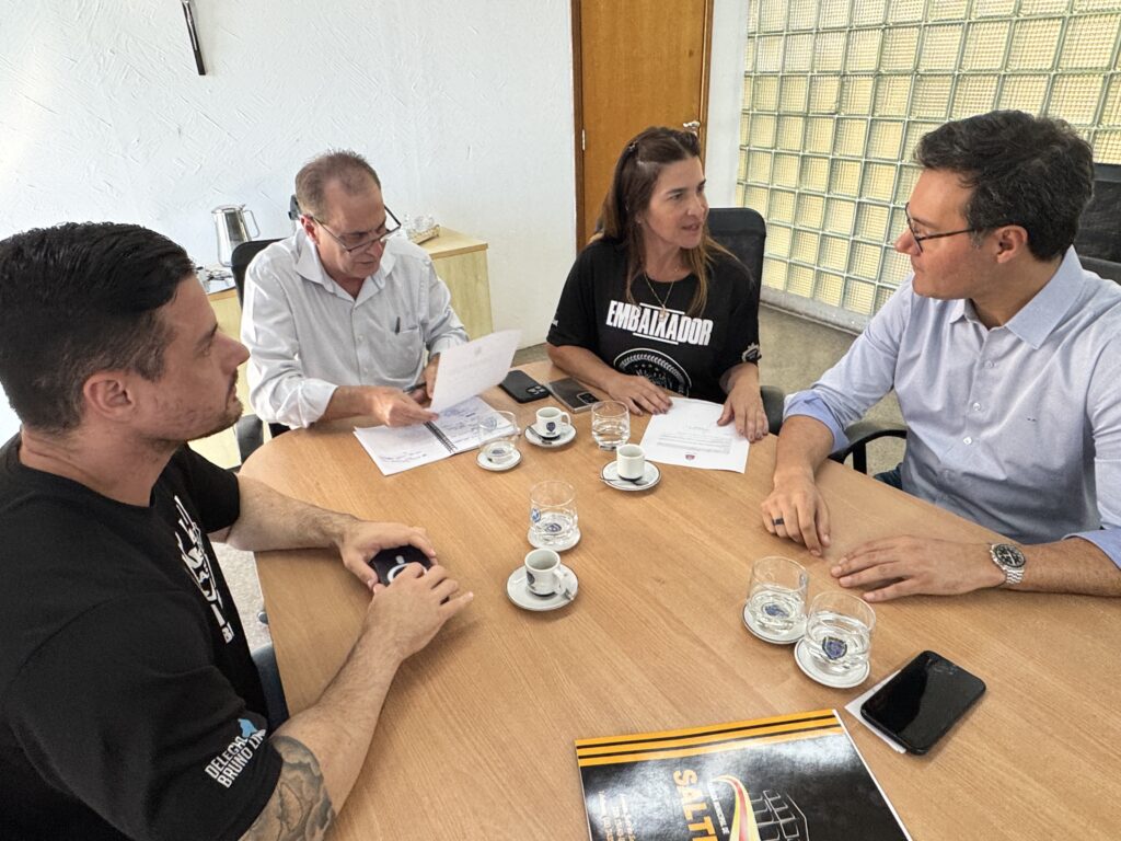 Na imagem aparece ao centro um senhor de óculos de camisa clara que está lendo uma folha, tendo ao seu lado um caderno. Também aparecem na foto uma mulher de blusa preta, com as mãos em cima de uma folha de papel, tendo ao seu lado um homem de óculos e do outro lado da mesa, aparece também um homem que veste uma camiseta preta com detalhes em branco. Estão reunidos em volta de uma mesa retangular de madeira clara, tendo em cima diversos copos e chicaras.