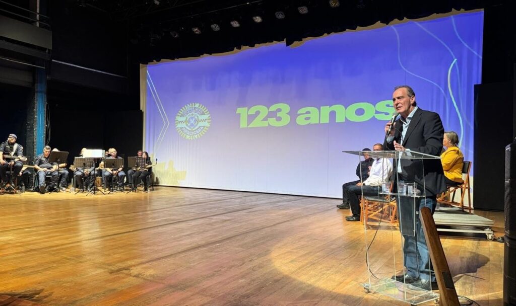 A imagem mostra um evento formal realizado em um palco, possivelmente em um teatro ou auditório. No centro da cena, um homem está falando em um púlpito transparente, segurando um microfone. Ele veste uma jaqueta escura, camisa azul clara e calça jeans. Ao fundo, há várias pessoas sentadas, entre elas uma mulher com uma jaqueta amarela. Também é possível ver um grupo musical sentado à esquerda, com instrumentos como saxofones e guitarras. No fundo do palco, uma grande tela exibe um fundo azul com linhas brancas onduladas e um logotipo circular com o texto "GUARDA CIVIL METROPOLITANA PIRACICABA". Em destaque, aparece o número "123 anos", indicando provavelmente a celebração do aniversário da instituição mencionada. O piso do palco é de madeira clara, refletindo a iluminação intensa que destaca o palestrante e a tela ao fundo. A imagem é capturada de um ângulo ligeiramente inferior, dando uma sensação de imponência ao evento.
