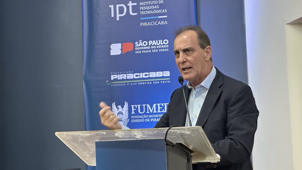 A imagem mostra um homem de meia-idade, com cabelo escuro e ralo, falando em um púlpito transparente. Ele veste um blazer azul escuro sobre uma camisa azul clara e está gesticulando com a mão direita enquanto fala, com expressão séria e concentrada. Atrás dele, há uma grande faixa azul com diversos logotipos e textos em branco e vermelho, indicando órgãos e instituições, entre eles "Instituto de Pesquisas Tecnológicas Piracicaba", "Governo do Estado de São Paulo", "Prefeitura Piracicaba" e "FUMEP - Fundação Municipal Ensino de Piracicaba". O cenário sugere que ele está em um evento oficial ou uma coletiva de imprensa, possivelmente falando sobre algum projeto ou iniciativa ligada à cidade de Piracicaba, no estado de São Paulo.