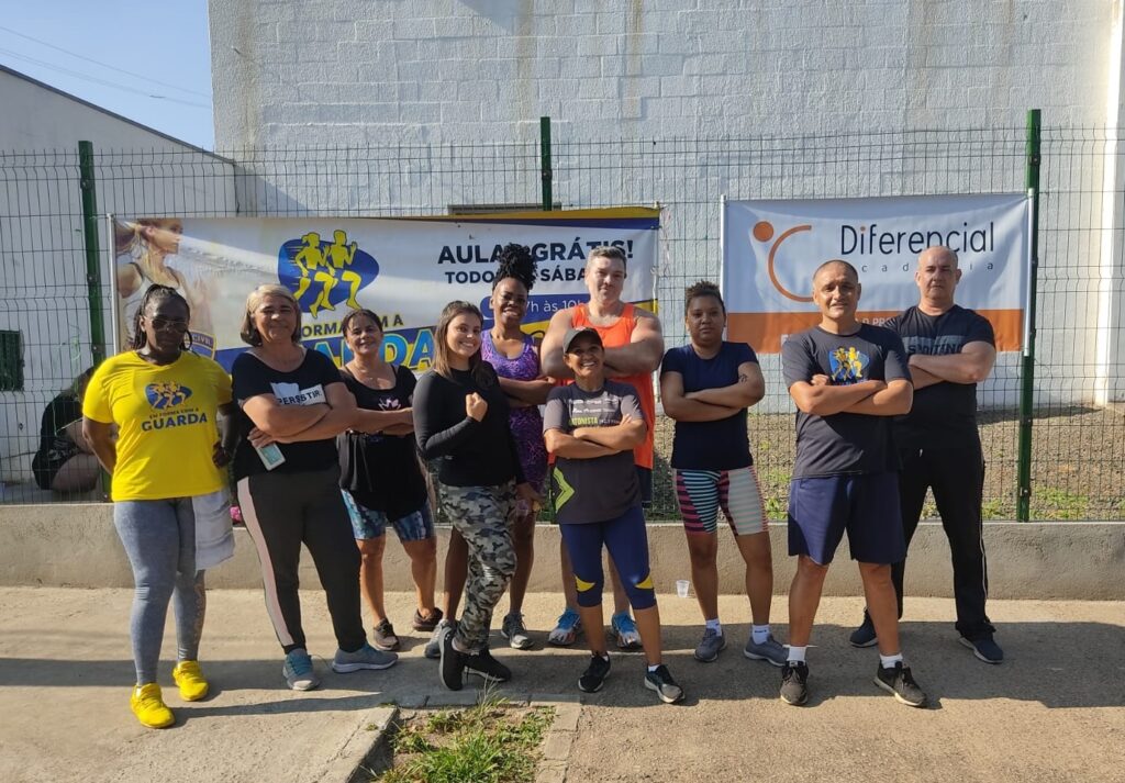 A imagem mostra um grupo de dez pessoas, cinco homens e cinco mulheres, posando para uma foto em grupo ao ar livre. Eles estão em frente a um muro de tijolos brancos e uma cerca verde de arame. Todos estão vestidos com roupas esportivas, como camisetas, leggings, shorts e tênis, sugerindo que participam de uma atividade física ou treino. Alguns estão com os braços cruzados e todos aparentam estar sorrindo ou com expressões confiantes. Atrás deles, penduradas na cerca, há duas faixas: uma delas oferece uma "AULA GRÁTIS! TODOS OS SÁBADOS, 7h às 10h" com a palavra "GUARDA" e um logo com figuras correndo; a outra apresenta o nome "Diferencial academia" e as palavras "PERSISTIR MARATONISTA EM FORMA COM A GUARDA". A foto foi tirada em um dia ensolarado, com sombras longas indicando que pode ser pela manhã. O chão é de concreto com um pequeno gramado na frente. O grupo está centralizado na imagem, aparecendo inteiro, em uma composição que destaca a união e o espírito esportivo do grupo.