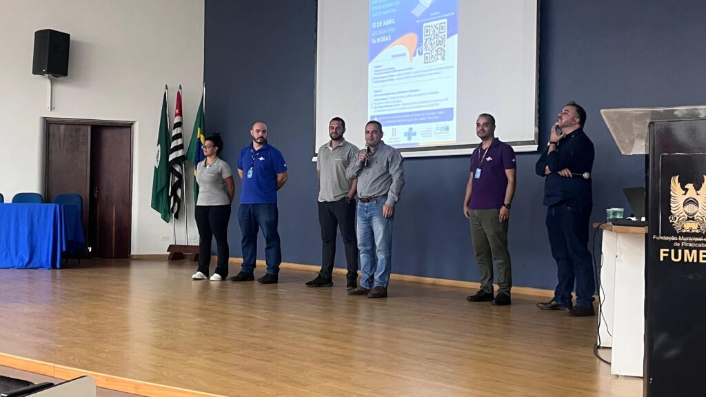 A imagem mostra um evento realizado em um auditório ou sala de conferências, com seis pessoas alinhadas no palco, todas de pé, algumas segurando microfones. Ao fundo, há uma parede azul escura com três bandeiras (duas delas são o estado de São Paulo e o Brasil) e um grande telão de projeção que exibe informações sobre o evento, incluindo a data "13 de abril", o dia da semana "segunda-feira" e o horário "14 horas". Também aparecem nomes e logos de organizações como SEBRAE, Fundação Municipal de Piracicaba e FUME. No palco, as pessoas estão vestidas de forma casual e semi-formal, com camisas pólo e calças. Algumas parecem estar falando ou prestes a falar, enquanto outras observam. A iluminação está focada no palco, destacando os participantes. À direita da imagem, há um púlpito com o logo da Fundação Municipal de Piracicaba. A cena sugere uma apresentação, palestra ou reunião oficial relacionada a temas de empreendedorismo ou cultura, dada a presença do SEBRAE e as referências visuais no telão, e parece ser um momento formal de exposição ou discussão.