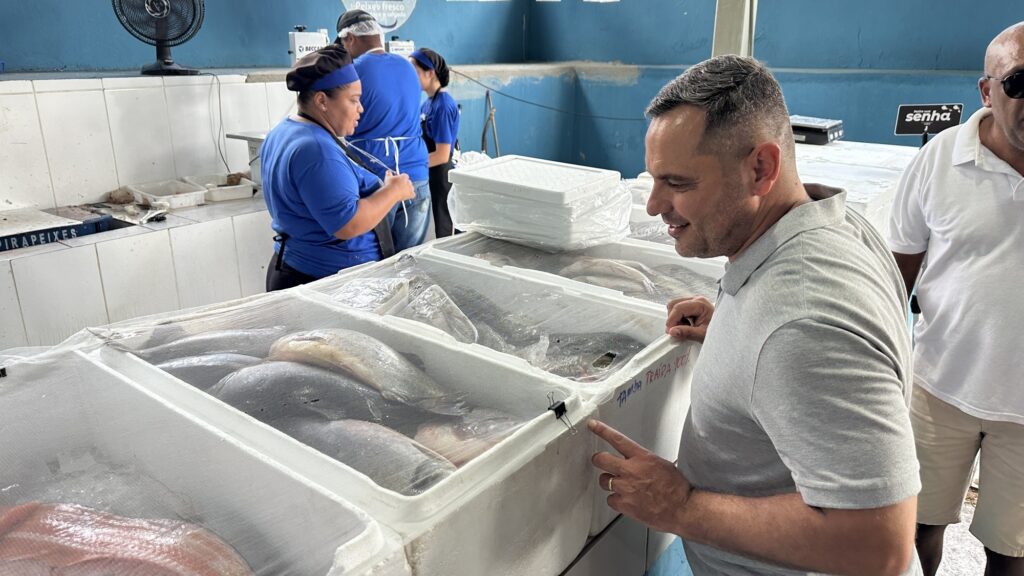 A imagem mostra um mercado de peixes, onde um homem está em destaque, sorrindo e apontando para uma caixa de isopor cheia de peixes frescos cobertos por uma tela plástica. Ele veste uma camisa pólo cinza. Ao fundo, há três funcionários do mercado, todos usando camisetas azuis e toucas, trabalhando atrás do balcão. Outro homem, de camiseta branca e óculos escuros, também aparece parcialmente. O ambiente é interno, com paredes azuis e balcões de azulejos brancos. Na frente, várias caixas de isopor com peixes estão alinhadas, e em uma delas está escrito à mão "TAMBA TRARA 100%". O local parece limpo e bem iluminado, transmitindo uma sensação de frescor e organização no comércio de peixes.