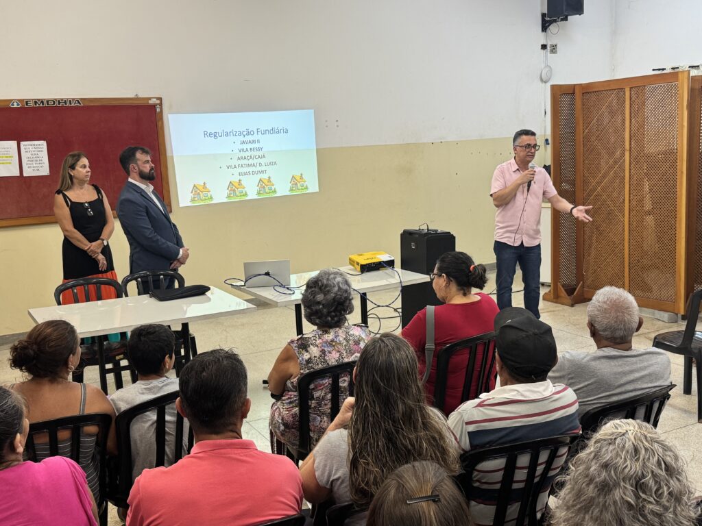 A imagem mostra uma reunião comunitária ou uma apresentação realizada em um ambiente interno bem iluminado, com paredes claras. À esquerda, uma mulher de regata preta e um homem de terno azul estão próximos a uma mesa branca, observando um homem que está falando para o público. O palestrante, um homem de meia-idade com óculos, veste uma camisa rosa de manga curta e calça jeans, segurando um microfone e gesticulando com a mão esquerda. Na parede ao fundo, um projetor exibe um slide com o título "Regularização Fundiária" e uma lista de localidades: JAVARI II, VILA BESSY, ARAÇÁ/CAJÁ, VILA FATIMA/D. LUIZA e ELIAS DUMIT, acompanhadas por ícones de pequenas casas. Em frente ao palestrante, várias pessoas estão sentadas em cadeiras de plástico pretas, assistindo à apresentação. A maioria das pessoas está com as costas para a câmera e vestem roupas casuais. Na parede à esquerda, há um quadro de avisos vermelho com o logotipo da EMDHIA e alguns comunicados. Também é possível ver equipamentos técnicos como um laptop, um projetor amarelo e uma caixa de som sobre mesas próximas ao centro da sala. A iluminação é clara e uniforme, indicando que a reunião ocorre durante o dia ou em um ambiente bem iluminado artificialmente.