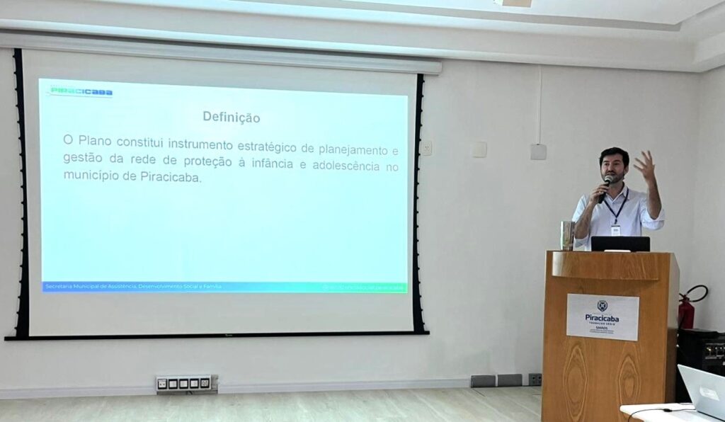 A imagem mostra um homem apresentando em uma sala de conferência ou reunião. Ele está em pé atrás de um púlpito de madeira, falando ao microfone, e parece estar explicando algo para uma audiência. O homem veste uma camisa azul clara e usa um crachá pendurado no pescoço. Ele está gesticulando com a mão direita enquanto fala. Ao fundo, há uma tela de projeção onde está exibido um slide com texto em português. O título do slide é "Definição" e o texto define o plano como um instrumento estratégico para o planejamento e gestão da rede de proteção à infância e adolescência no município de Piracicaba. Na parte inferior do slide, há menção à Secretaria Municipal de Assistência, Desenvolvimento Social e Família. No púlpito, há um logotipo e o nome "PIRACICABA" e a frase "TRABALHO SÉRIO". A sala é clara, com paredes claras e tomadas visíveis na parede ao fundo. A imagem está bem iluminada e focada, mostrando claramente o apresentador e o conteúdo do slide.