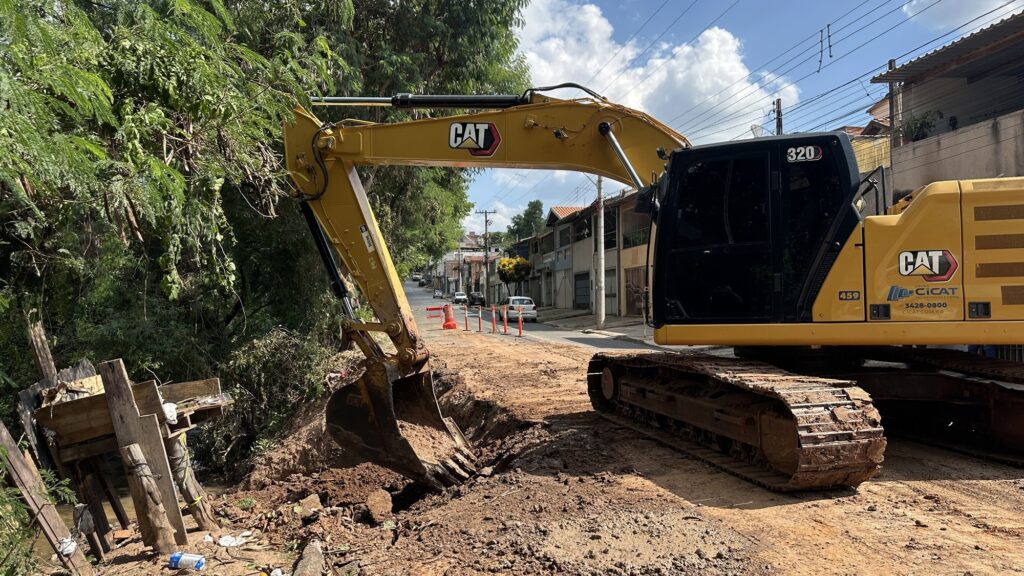 A imagem mostra uma escavadeira hidráulica amarela da marca CAT, modelo 320, em operação em uma rua de terra. A máquina está escavando uma vala ao longo da estrada, com o braço da escavadeira estendido e a caçamba cheia de terra. A escavadeira está apoiada sobre esteiras que estão cobertas de lama, indicando que o terreno está úmido ou recentemente trabalhado. Ao redor da escavadeira, há vegetação densa, com árvores e arbustos, principalmente à esquerda da imagem. Ao fundo, do outro lado da rua, há casas e edifícios residenciais, sugerindo que a obra está ocorrendo em uma área urbana ou semiurbana. No chão, podem ser vistos cones de sinalização laranja, delimitando a área de trabalho. Na lateral da escavadeira, está a inscrição “GRUPO CICAT PAVIMENTAÇÃO E OBRAS”, além do telefone de contato (11) 3428-0800 e o site cicat.com.br, indicando a empresa responsável pela obra. O número “459” também está visível na máquina. O céu está parcialmente nublado, com nuvens brancas e céu azul, indicando um dia claro. A iluminação natural destaca a cor amarela vibrante da escavadeira e o contraste com a terra e a vegetação. A perspectiva da foto é próxima e ligeiramente inclinada para cima, dando destaque à escavadeira como o elemento principal da cena. No geral, a imagem representa uma obra de construção ou manutenção viária, com a escavadeira realizando uma escavação para provavelmente instalar ou reparar alguma infraestrutura no local.