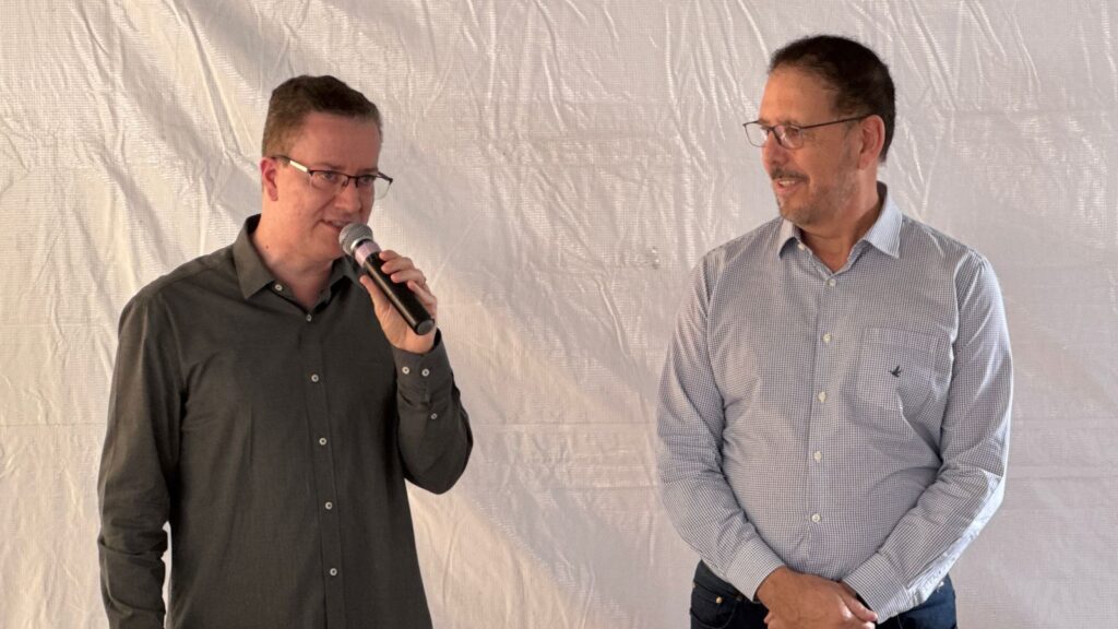 A imagem mostra um homem de óculos de camisa escura fala ao microfone, tendo ao seu lado um senhor de óculos e camiseta clara listrada. Ao fundo tem uma parede com uma lona branca. Tudo indica ser um evento de inauguração.