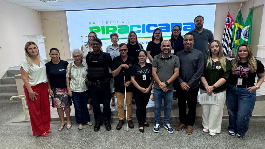 A imagem mostra um grupo de 13 pessoas posando para uma foto em um ambiente interno, possivelmente uma sala de eventos ou auditório de um prédio público. O grupo está distribuído em duas fileiras: algumas pessoas estão em pé atrás e outras estão sentadas ou agachadas na frente. Ao fundo, há uma grande tela digital que exibe o texto "PREFEITURA PIRACICABA" em letras grandes, com um design colorido e moderno. Ao lado da tela, há três bandeiras: a bandeira do estado de São Paulo, a bandeira do Brasil e outra bandeira verde com brasão, provavelmente do município de Piracicaba. As pessoas na imagem são diversas em termos de idade e gênero, vestindo roupas que vão do casual ao mais formal. Entre eles, uma pessoa segura uma bengala branca, indicando que é deficiente visual. A iluminação do local é clara e uniforme, típica de ambientes internos bem iluminados. À esquerda, é possível ver uma escada, sugerindo que o local é uma área pública ou administrativa. No geral, a foto parece ter sido tirada para registrar um evento oficial ou uma reunião promovida pela Prefeitura de Piracicaba, com um grupo que pode incluir servidores, convidados ou participantes de algum programa ou projeto. A atmosfera é formal, porém descontraída, com algumas pessoas sorrindo para a câmera.