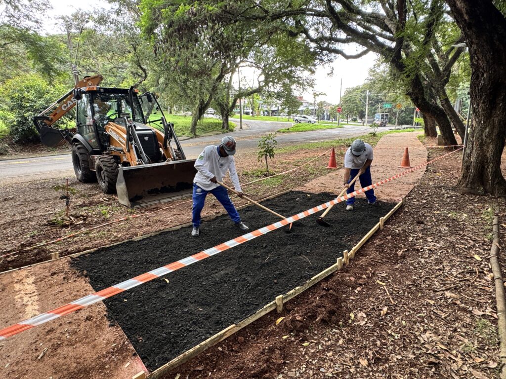 A imagem mostra uma cena de construção ao ar livre, possivelmente em um parque ou à beira de uma estrada. Dois trabalhadores estão no centro da imagem, ambos usando capacetes de segurança e roupas de trabalho, enquanto espalham e nivelam asfalto recém-colocado com ancinhos. O asfalto está contido dentro de uma moldura feita com tábuas de madeira e cercado por uma fita de segurança laranja e branca, indicando área de obra. À esquerda, há uma retroescavadeira laranja da marca CASE, com a pá levantada. O chão ao redor é de terra avermelhada e há algumas folhas espalhadas, sugerindo um ambiente natural. Ao fundo, há várias árvores com folhagem verde e uma estrada asfaltada onde passam alguns veículos, incluindo um caminhão branco. A iluminação é suave, indicando que pode ser um dia nublado ou um momento próximo ao entardecer. A cena transmite um momento de trabalho manual na construção ou reparo de uma via, mostrando a interação entre o esforço humano e o maquinário pesado em um ambiente natural.