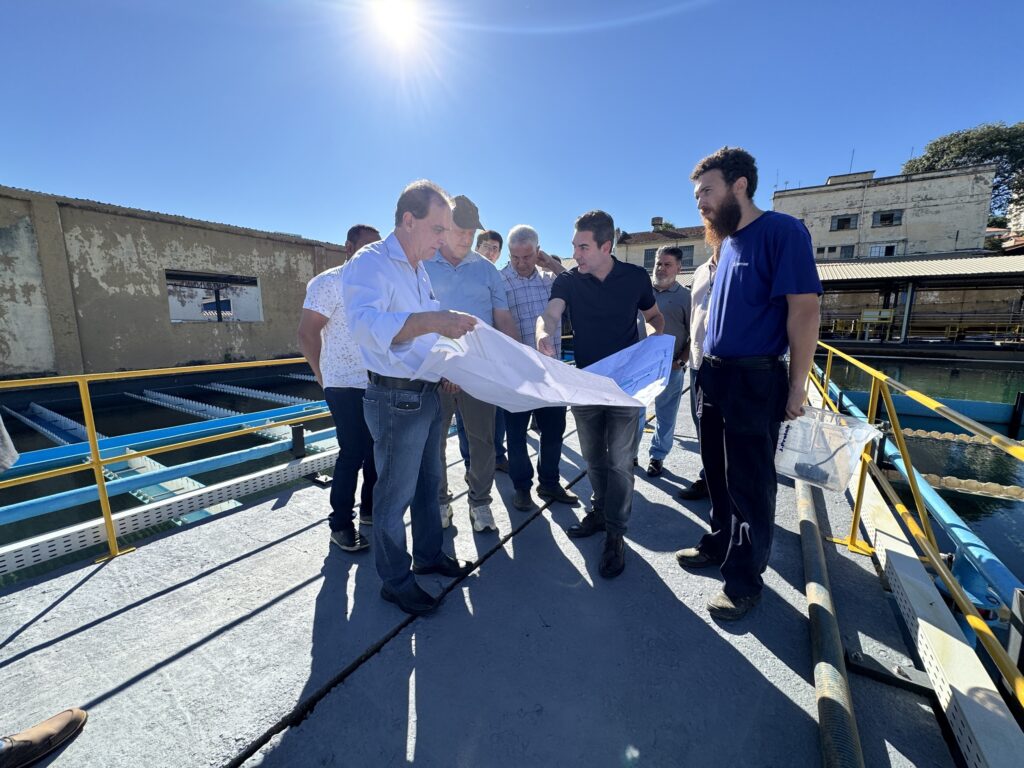 A imagem mostra um grupo de nove homens reunidos ao ar livre, em um ambiente industrial que parece ser uma estação de tratamento de água. Dois homens estão em destaque no primeiro plano, segurando e analisando um grande documento branco, possivelmente plantas ou projetos. Um deles, vestido com uma camisa azul clara e jeans, aponta para o documento, enquanto o outro, com uma camisa preta e calça escura, observa atentamente. Os demais homens estão ao redor, observando ou ouvindo a conversa. O cenário é uma plataforma com corrimãos azuis e amarelos, acima de tanques ou bacias de água. Ao fundo, há um prédio de concreto desgastado com uma grande janela aberta e algumas máquinas industriais. O céu está claro e azul, com o sol brilhando intensamente, criando sombras fortes no chão. A imagem transmite uma reunião profissional, provavelmente uma inspeção ou discussão técnica relacionada à infraestrutura ou engenharia.