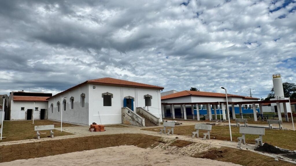 A imagem mostra uma área pública ao ar livre, provavelmente uma praça ou um centro comunitário em uma pequena cidade ou zona rural. No centro da cena, há um conjunto de edifícios brancos de um andar com telhados de telha cerâmica vermelha. Um dos prédios possui uma escadaria com a palavra "CAPA" grafitada na lateral. No chão, há várias estruturas que parecem bancos de concreto espalhados, e um monte de tijolos vermelhos empilhados próximo ao prédio central, sugerindo que a área está em processo de reforma ou construção. Ao fundo, à direita, é possível ver uma torre de água ou um silo branco. O céu está muito nublado, com nuvens escuras que dão um aspecto dramático à cena. O chão é uma mistura de terra, areia e algumas áreas com vegetação rasteira. Não há pessoas visíveis na imagem, e a iluminação é difusa devido ao céu encoberto. No geral, a imagem transmite uma sensação de tranquilidade e simplicidade, típica de uma pequena comunidade, com um toque de atividade recente ou em andamento, devido à presença dos tijolos e ao estado do local.