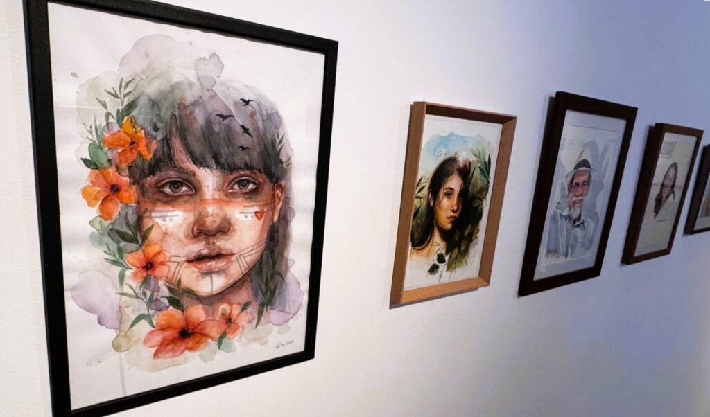 A imagem mostra uma exposição de arte com várias pinturas em aquarela penduradas em uma parede branca. A pintura em destaque, a mais próxima e maior, retrata uma jovem mulher com cabelo escuro, olhos expressivos e pintura tribal no rosto, incluindo marcas brancas nas bochechas e um coração no lado direito. Ela está adornada com flores laranja e folhagens verdes ao redor, e há silhuetas de pássaros voando acima de sua cabeça. O estilo é suave e expressivo, com pinceladas visíveis e texturas típicas da aquarela. Ao lado dessa obra principal, há outras pinturas menores em sequência, formando uma fila que se estende para o fundo da imagem. A segunda pintura mostra outra jovem mulher com cabelo castanho longo, com fundo de céu azul e vegetação. A terceira retrata um homem mais velho, de barba branca e chapéu, também em aquarela. As molduras variam em cor e material, desde preto até madeira clara e escura. A iluminação é suave, provavelmente proveniente de luzes no teto, destacando os detalhes e texturas das obras. A exposição transmite uma sensação de cuidado artístico e uma estética natural e etérea.