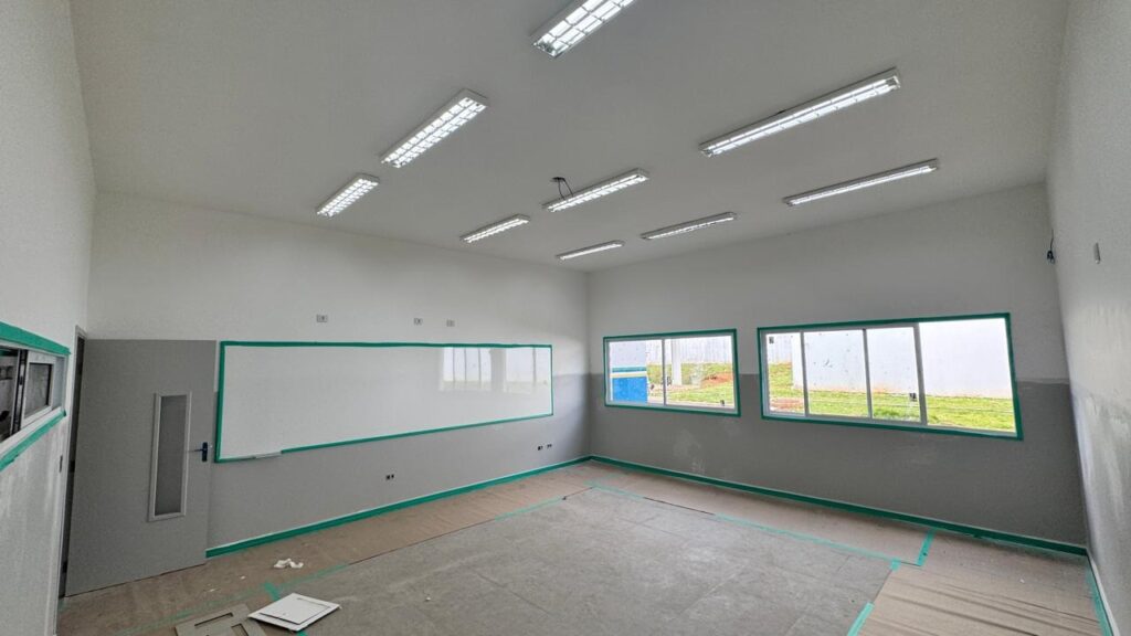 A imagem mostra uma sala de aula vazia, recém-construída ou em fase final de reforma. As paredes são pintadas em tons neutros, com a parte superior branca e a inferior cinza, e há faixas de fita verde de pintura ao longo das bordas, indicando que o acabamento ainda está em andamento. No lado esquerdo da sala, há um grande quadro branco com moldura verde fixado na parede. À esquerda do quadro, há uma porta cinza com uma pequena janela vertical de vidro. No lado direito da sala, há duas janelas grandes que permitem a entrada de luz natural, mostrando uma área externa com grama e um muro branco. O teto é branco e possui várias luminárias retangulares embutidas que fornecem iluminação uniforme. O piso parece estar coberto por um material protetor, possivelmente papel ou plástico, sugerindo que o espaço ainda está sendo preparado para uso. A sala está limpa, organizada e sem móveis.