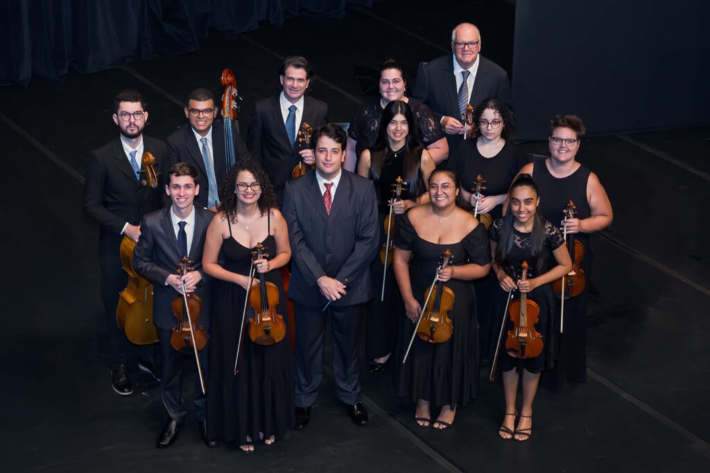 A imagem mostra uma orquestra de câmara pequena composta por treze músicos, posando juntos em um palco escuro. Eles estão organizados em três fileiras. Na fileira da frente, no centro, está um maestro vestido com um terno escuro listrado e gravata vermelha, segurando uma batuta. Ao redor dele, há músicos tocando instrumentos de corda, como violinos e violas, vestidos majoritariamente com roupas pretas ou ternos escuros. Na fileira do meio, há cinco músicos, incluindo homens e mulheres, também com instrumentos de corda, e na fileira de trás, quatro pessoas, entre elas um homem mais velho com terno e gravata. A iluminação é clara e uniforme, destacando os músicos contra o fundo escuro do palco, que possui cortinas pretas à esquerda. A atmosfera da foto é formal e profissional, transmitindo um momento de celebração ou apresentação.