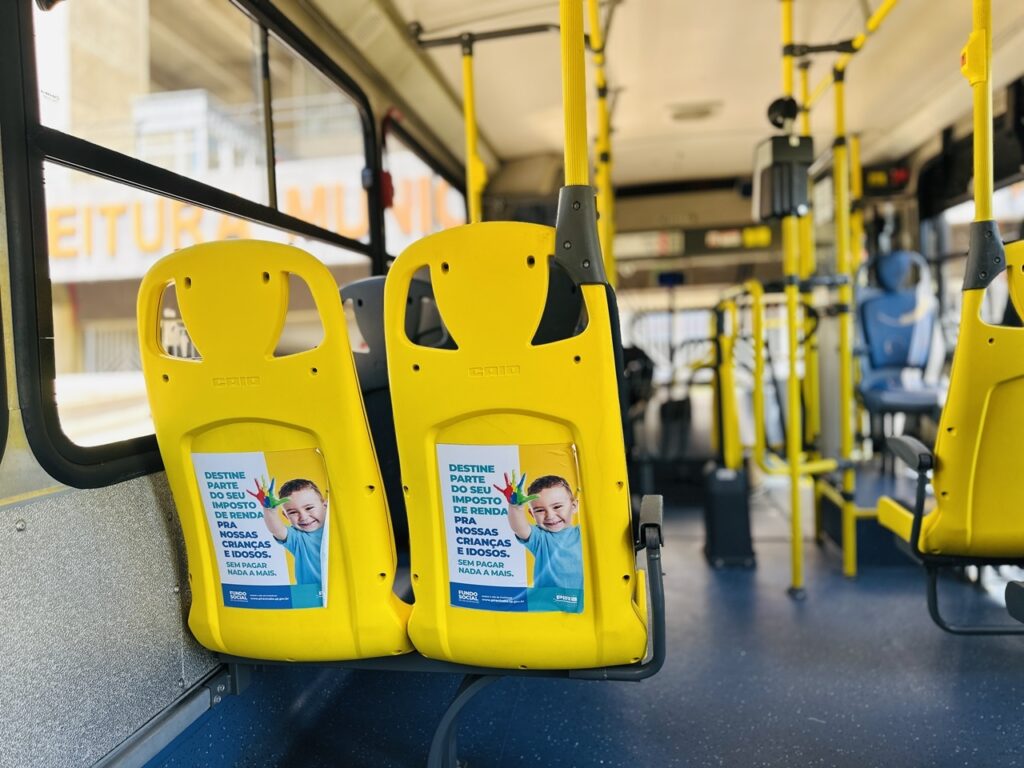 A imagem mostra o interior de um ônibus público, com foco em dois assentos amarelos lado a lado na parte dianteira do veículo. Cada assento possui um adesivo colado no encosto, contendo uma mensagem em português que incentiva a destinação de parte do imposto de renda para ajudar crianças e idosos, sem custo adicional para o contribuinte. O adesivo apresenta a imagem de uma criança sorridente com mãos pintadas de várias cores, simbolizando apoio e solidariedade. O interior do ônibus é predominantemente amarelo, com barras de apoio também na cor amarela, e o piso é azul. Ao fundo, é possível ver mais assentos azuis e janelas que mostram a paisagem urbana do lado de fora, incluindo um prédio com parte da palavra "EITUR" visível, provavelmente parte da palavra "PREFEITURA". A iluminação é clara, sugerindo que a foto foi tirada durante o dia. A composição destaca a campanha social presente nos adesivos, enfatizando a importância da contribuição para causas sociais por meio do imposto de renda.